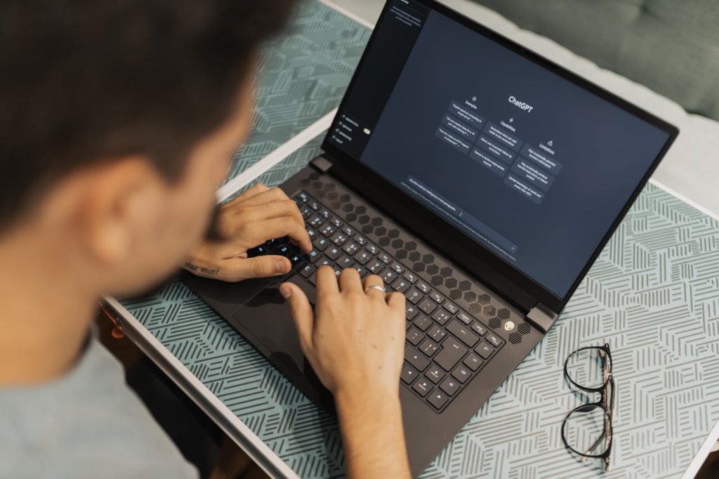 man-using-chatgpt-16094046 A man working on a laptop with AI software open on the screen, wearing eyeglasses.